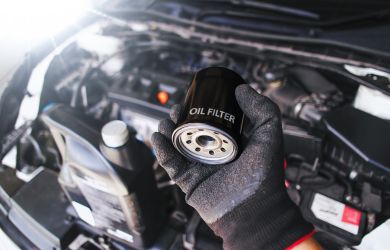 A mechanic wearing gloves holding a black oil filter in front of an open car engine bay, with a bottle of engine oil in the background.