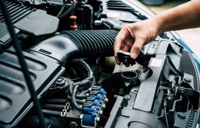 A person’s hand removing the radiator cap of a car engine for inspection, with various engine components and hoses visible under the hood.