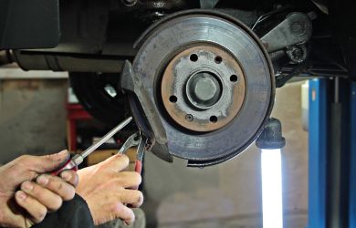 A mechanic using pliers to work on a vehicle's exposed brake disc and caliper assembly in a garage setting, with a bright work light illuminating the area.