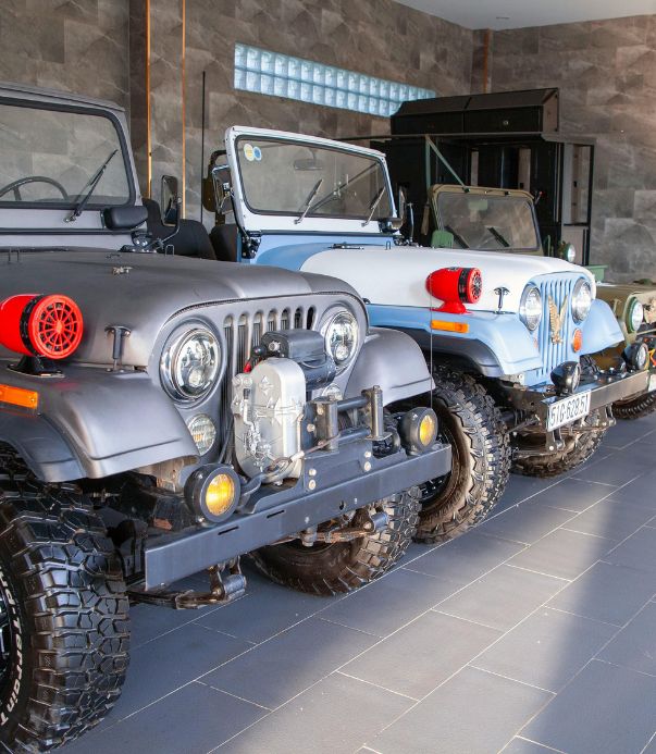 A row of three classic off-road Jeep vehicles parked indoors. The first Jeep is matte black with a front-mounted winch and red accessories.